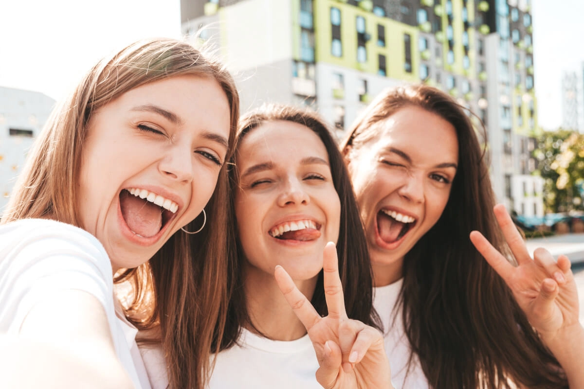 Drei junge Frauen machen ein fröhliches Selfie im Sommer vor modernen Stadtgebäuden.