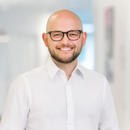 Porträt eines lächelnden Mannes mit Brille im modernen, hellen Büro-Umfeld.